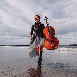 Woman in grey long dress and black short jacket running along the beach holding a cello in her left hand. Landscape shows grey skies and sea shore just behind her.
