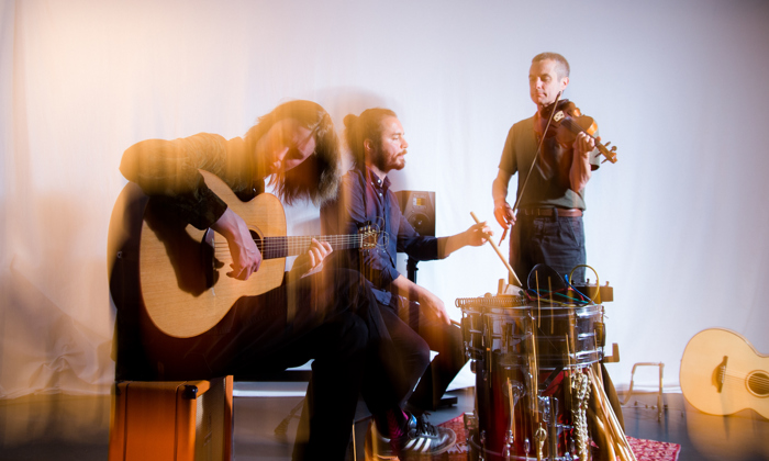 Three musicians from the band Coras Trio, one is sitting playing an acoustic guitar, another the drums and the third is standing up playing the violin.